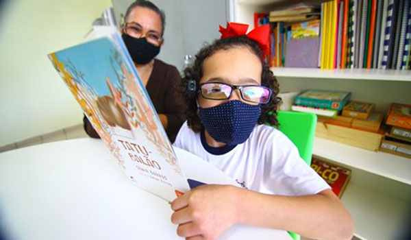 Yasmin, de 6 anos, cega, está segurando um livro e, utilizando seu OrCam MyEye, está usando-o para leitura completa do material.