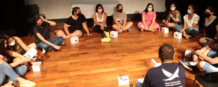 Em um palco de teatro, diversas pessoas estão sentadas em um círculo, observando o treinamento de um profissional da Mais Autonomia - Mais Autonomia