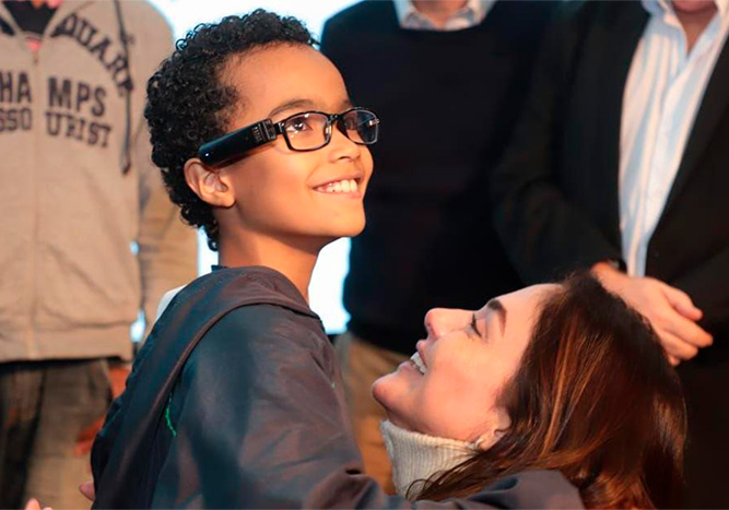 Menino usando o Orcam MyEye ao lado de sua mãe